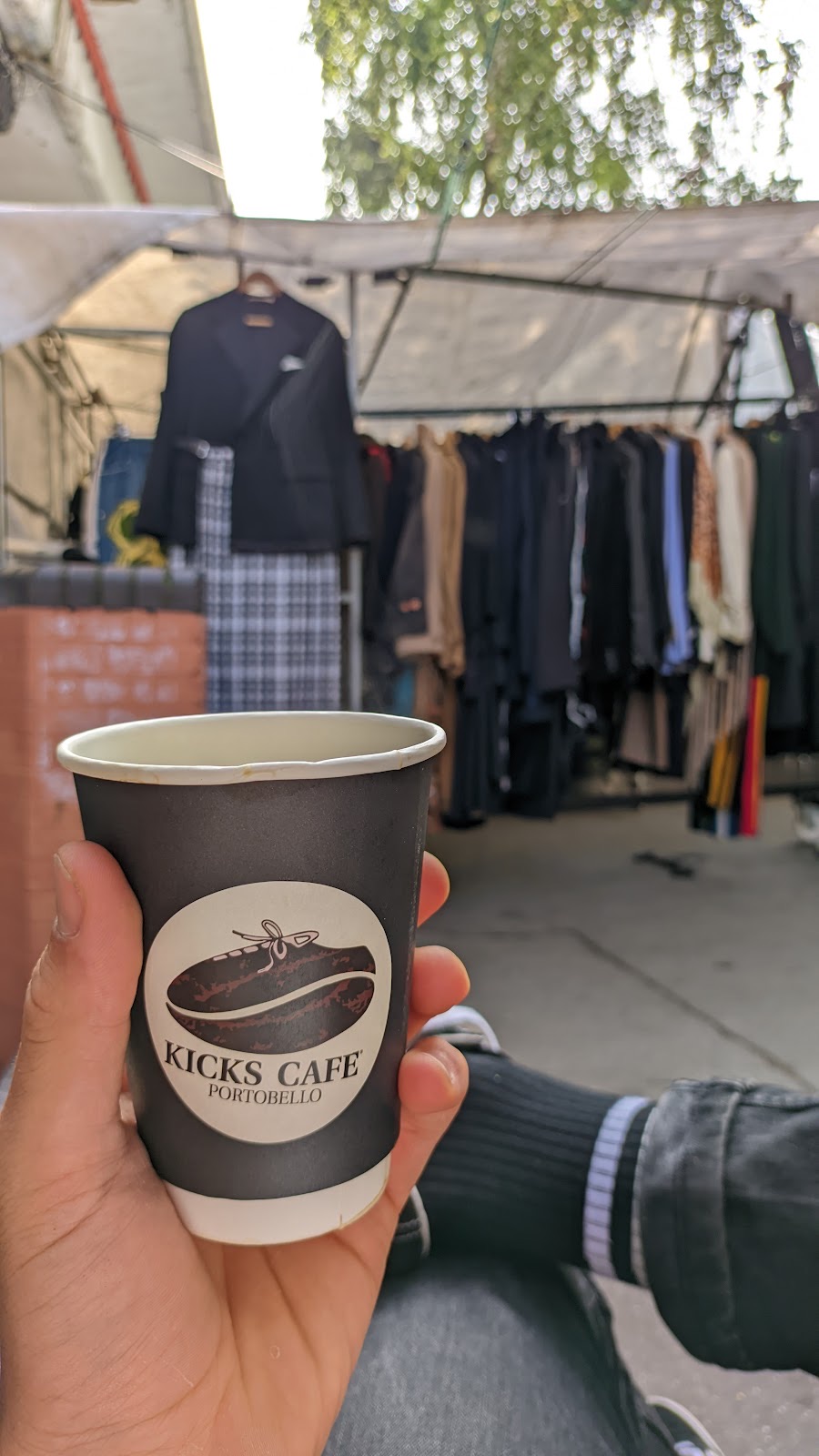 A hand holding a Kicks Cafe takeaway coffee cup on Portobello Road, the market stalls behind